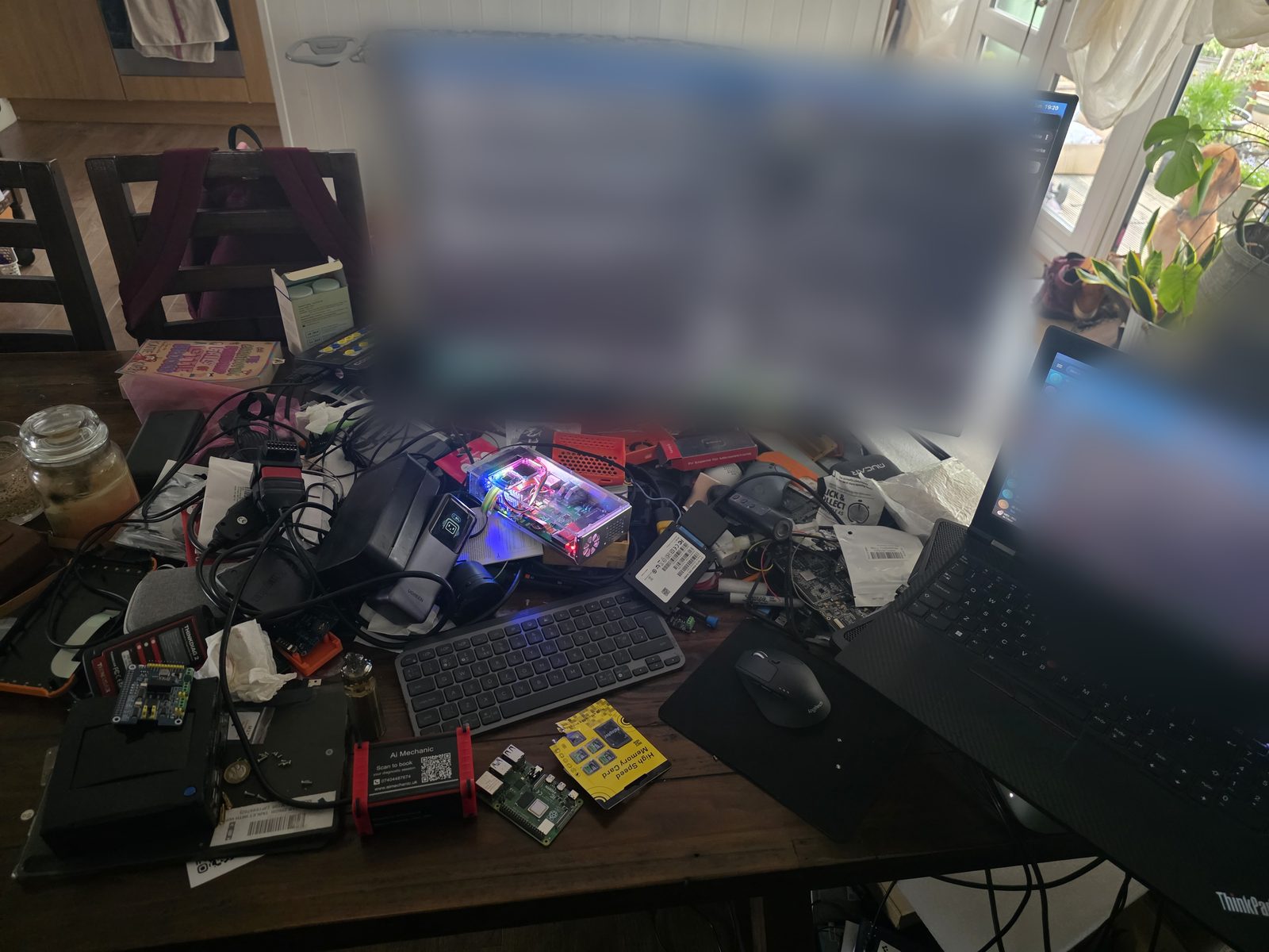 John's actual workspace: half a dining table covered in OBD dongles, ECUs, an open PC with a glowing circuit board, diagnostic tools, two computers, and a candle jar holding the line for domestic life.